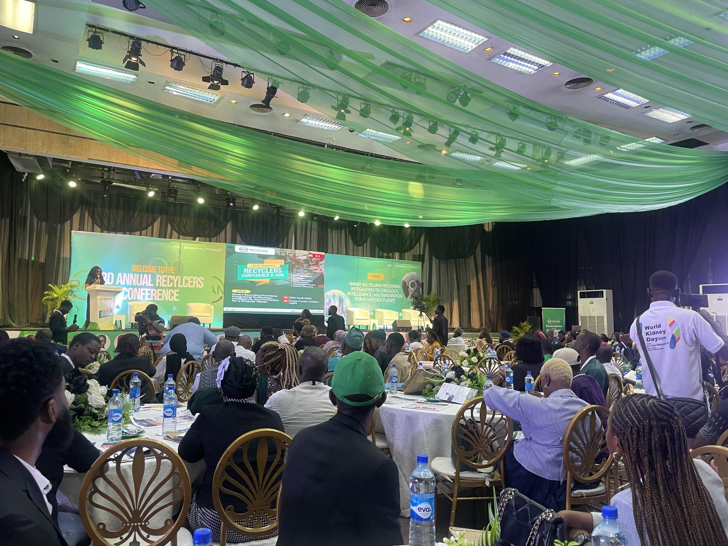 The conference hall seen from the audience, with delegates seated at round tables under green drapes and the stage screens reading Welcome to the 3rd Annual Recyclers Conference.