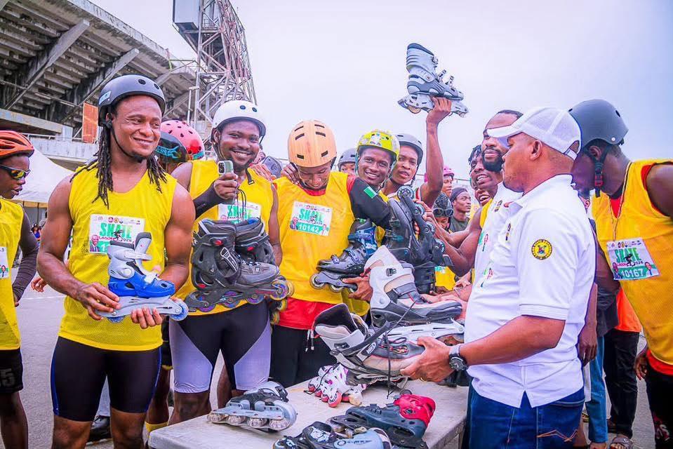 Skaters in yellow event bibs and helmets holding pairs of inline skates, with a man in a white polo shirt holding another pair in front of them and more laid out on a table.