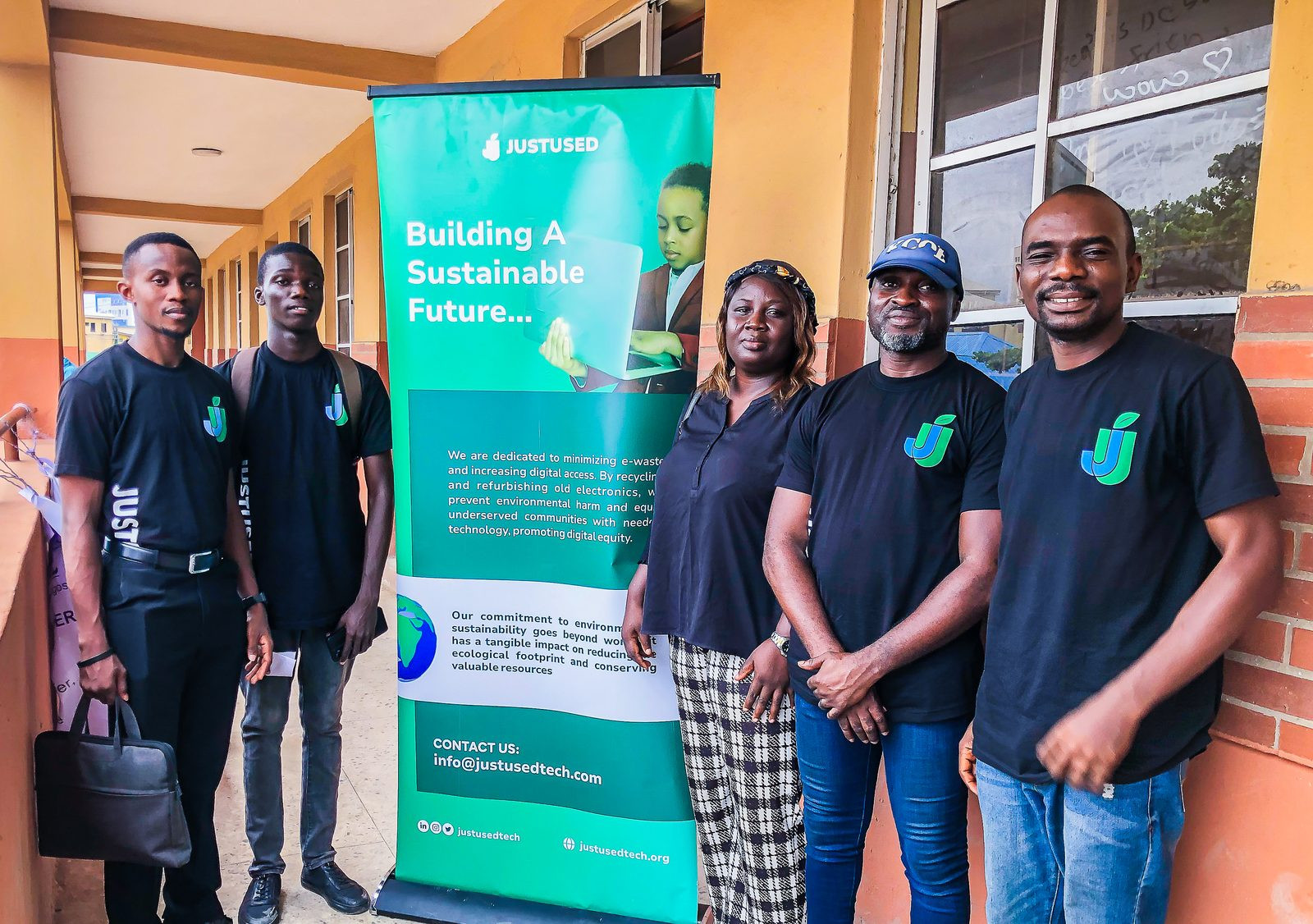 Five team members in JustUsedTech shirts standing beside a Building A Sustainable Future banner on a school walkway. (8 schools in Lagos State)