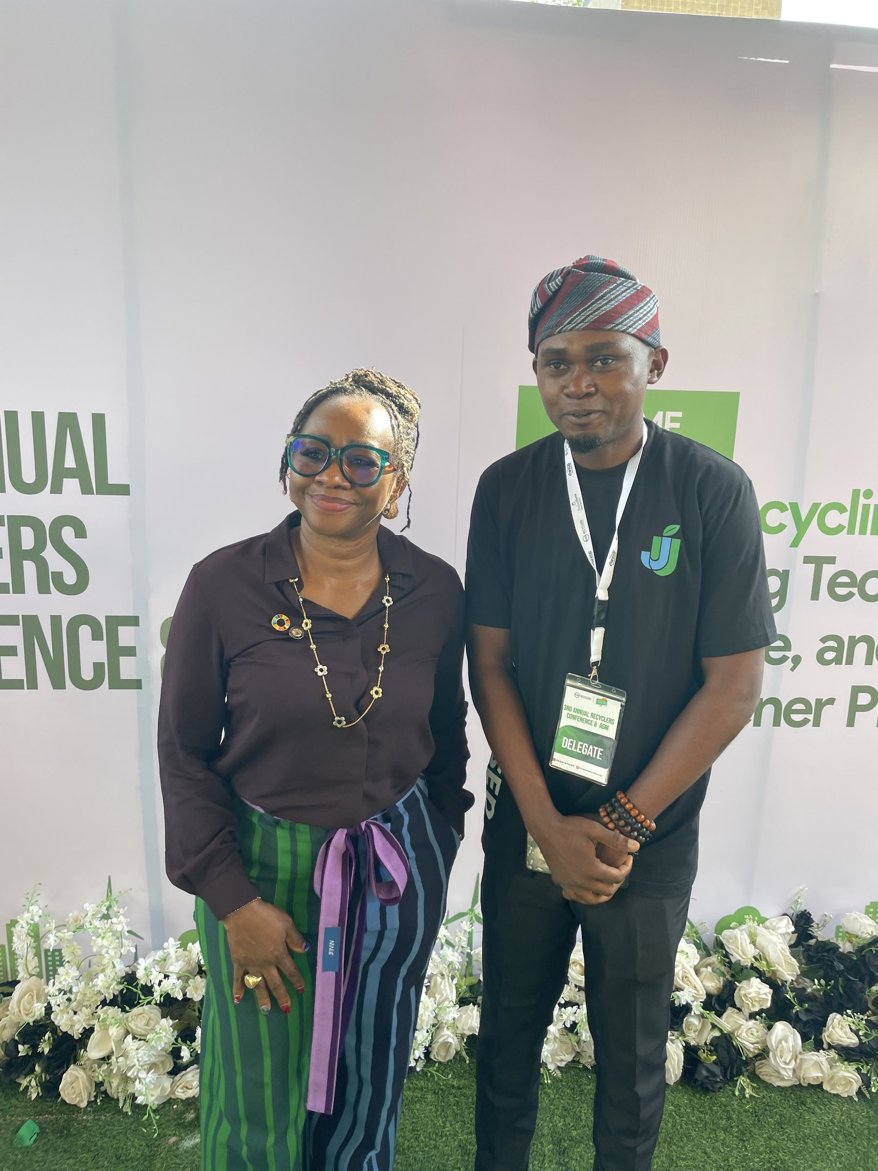A JustUsedTech team member in a JUSTUSED shirt and a delegate badge standing with another attendee in front of the conference backdrop.