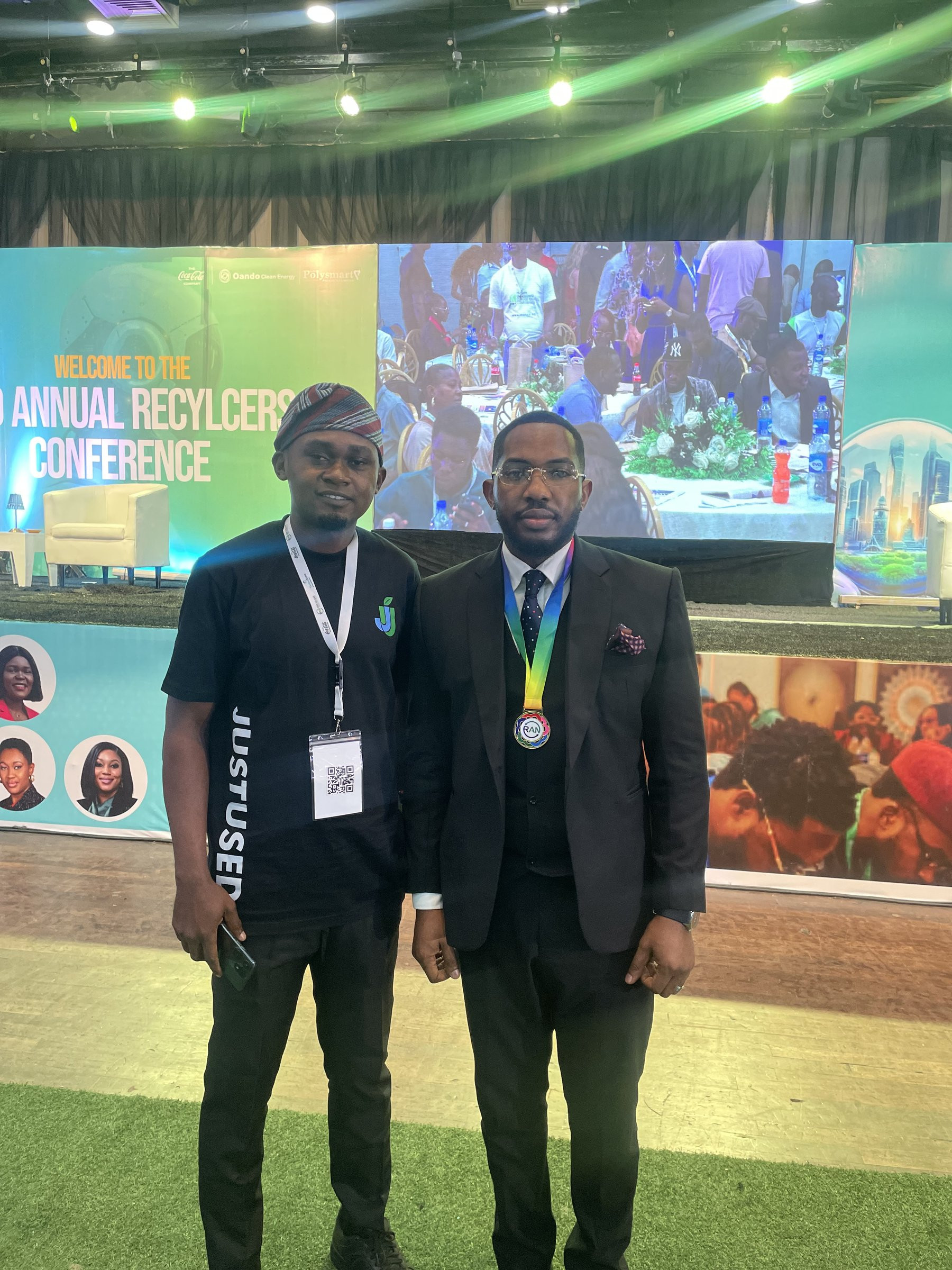 A JustUsedTech team member in a JUSTUSED shirt standing with another attendee in a suit and medal in front of the conference stage.