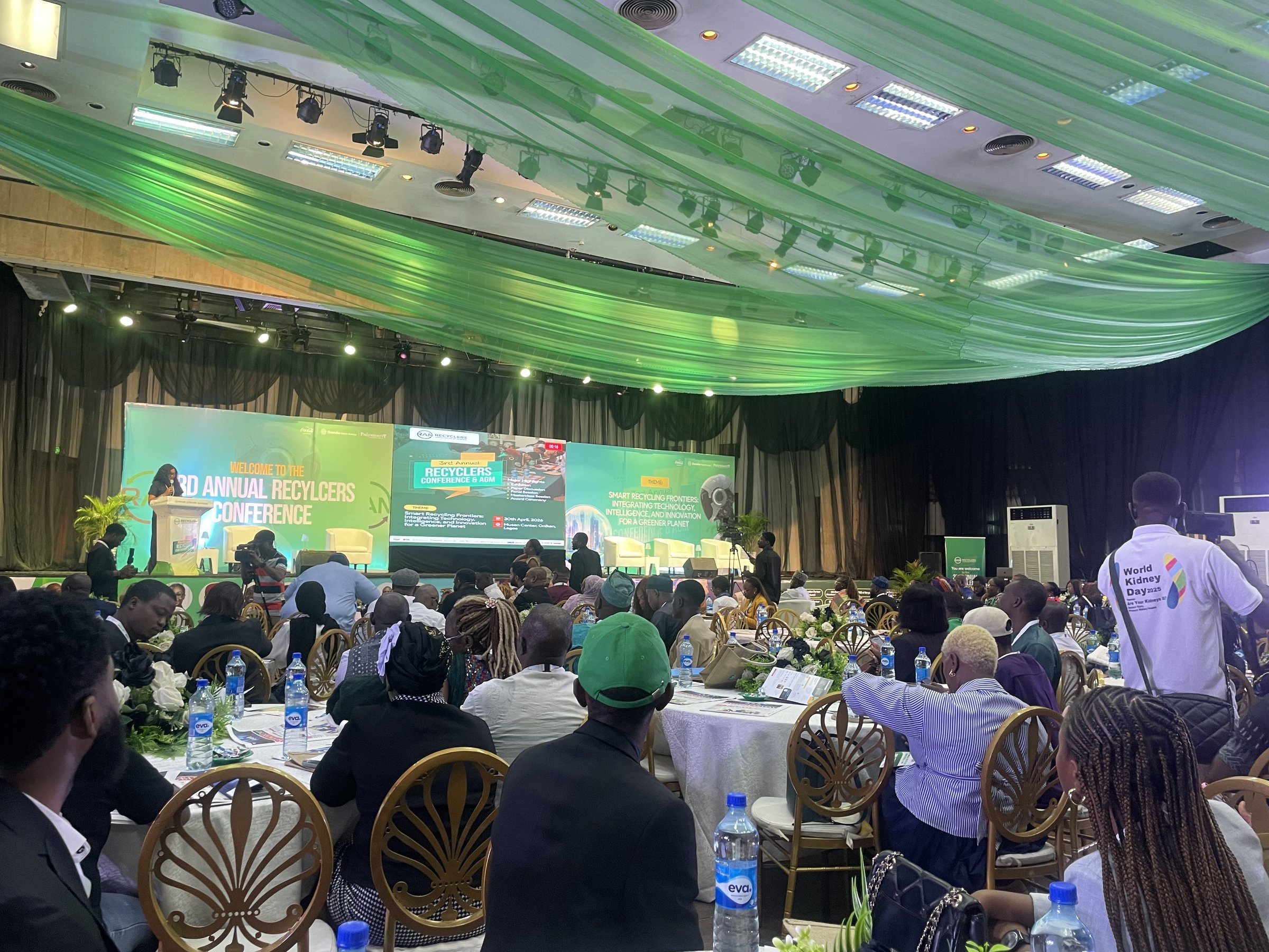 The conference hall seen from the audience, with delegates seated at round tables under green drapes and the stage screens reading Welcome to the 3rd Annual Recyclers Conference.