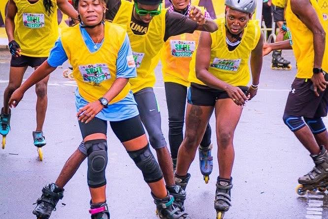 Four skaters in yellow event bibs, helmets, and knee pads skating together along a road.