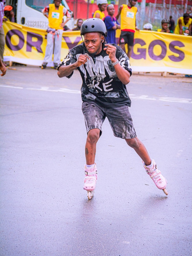 A skater in a helmet and printed shirt mid-turn on a road, in front of a yellow Sporty Lagos banner.