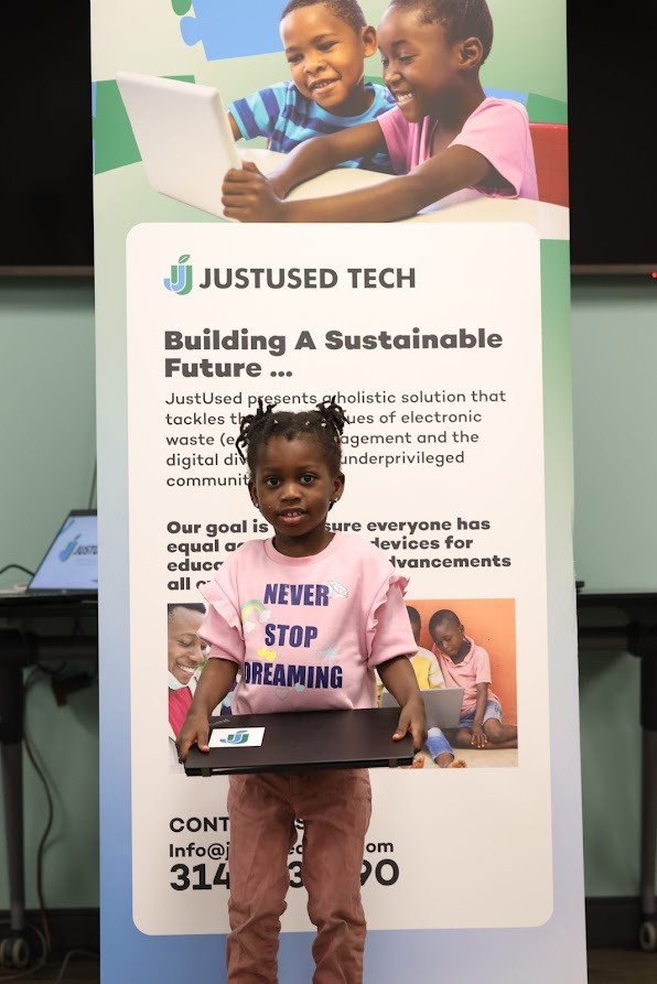 A child holding a laptop, standing beside a JustUsedTech roll-up banner.