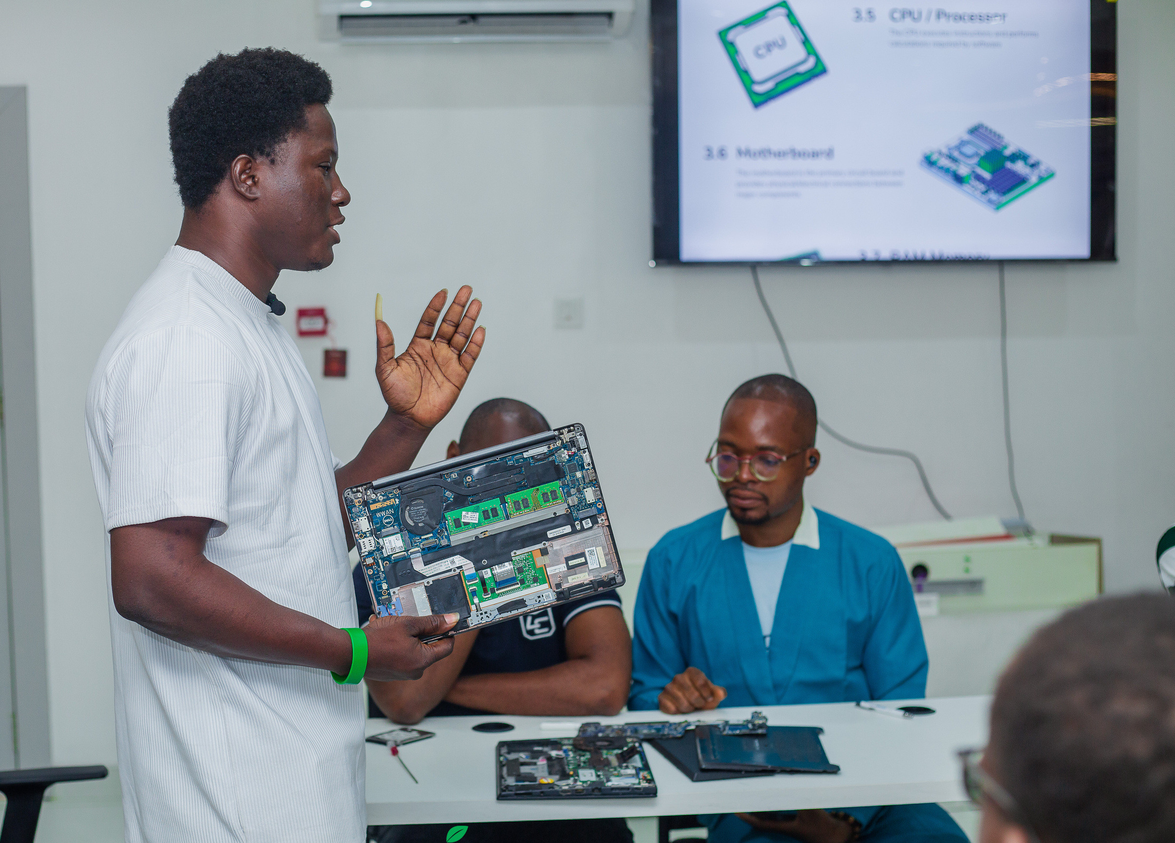 A facilitator holding an opened laptop chassis up to the room, with the component slide on the screen behind.
