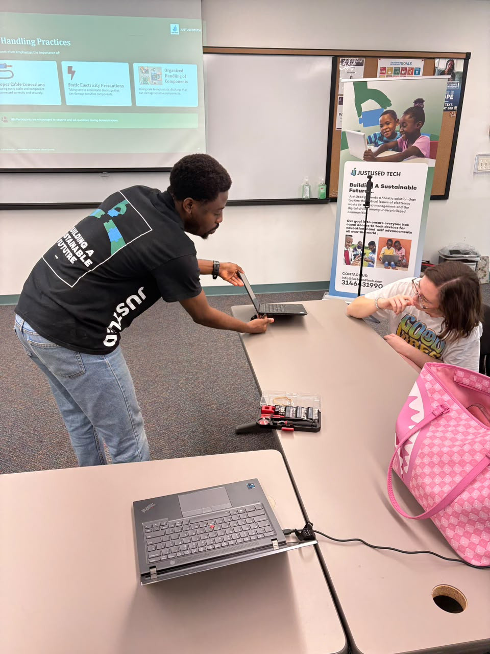 A team member leaning across a table to show a laptop to a young participant, beside a JustUsedTech poster.