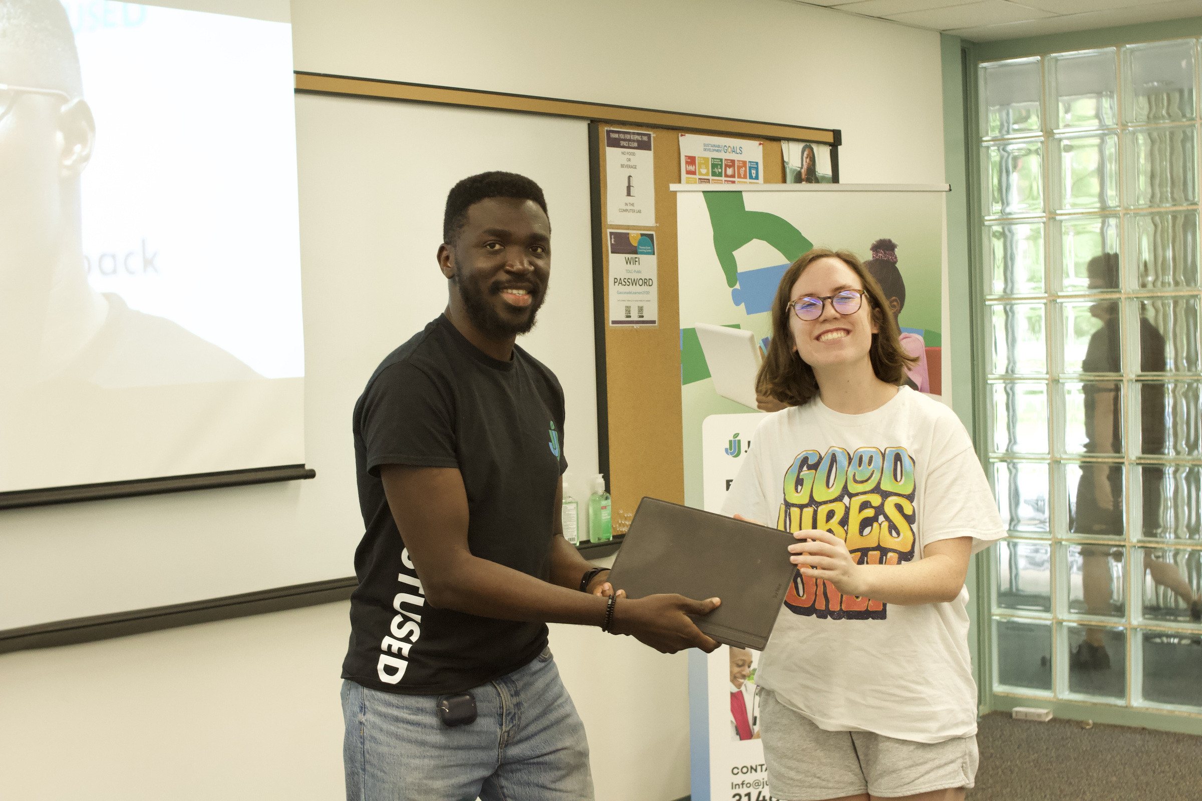A team member handing a laptop to a participant beside a JustUsedTech banner.