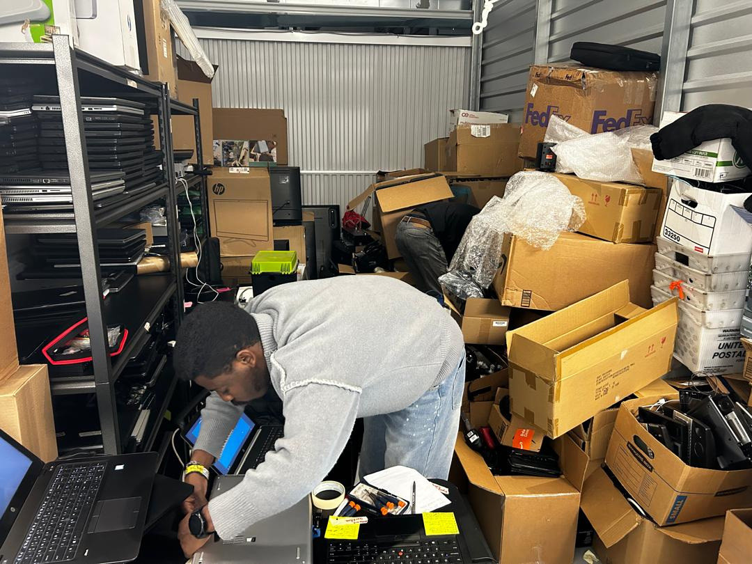 A person sorting laptops in a storage area stacked with cartons and boxed equipment.