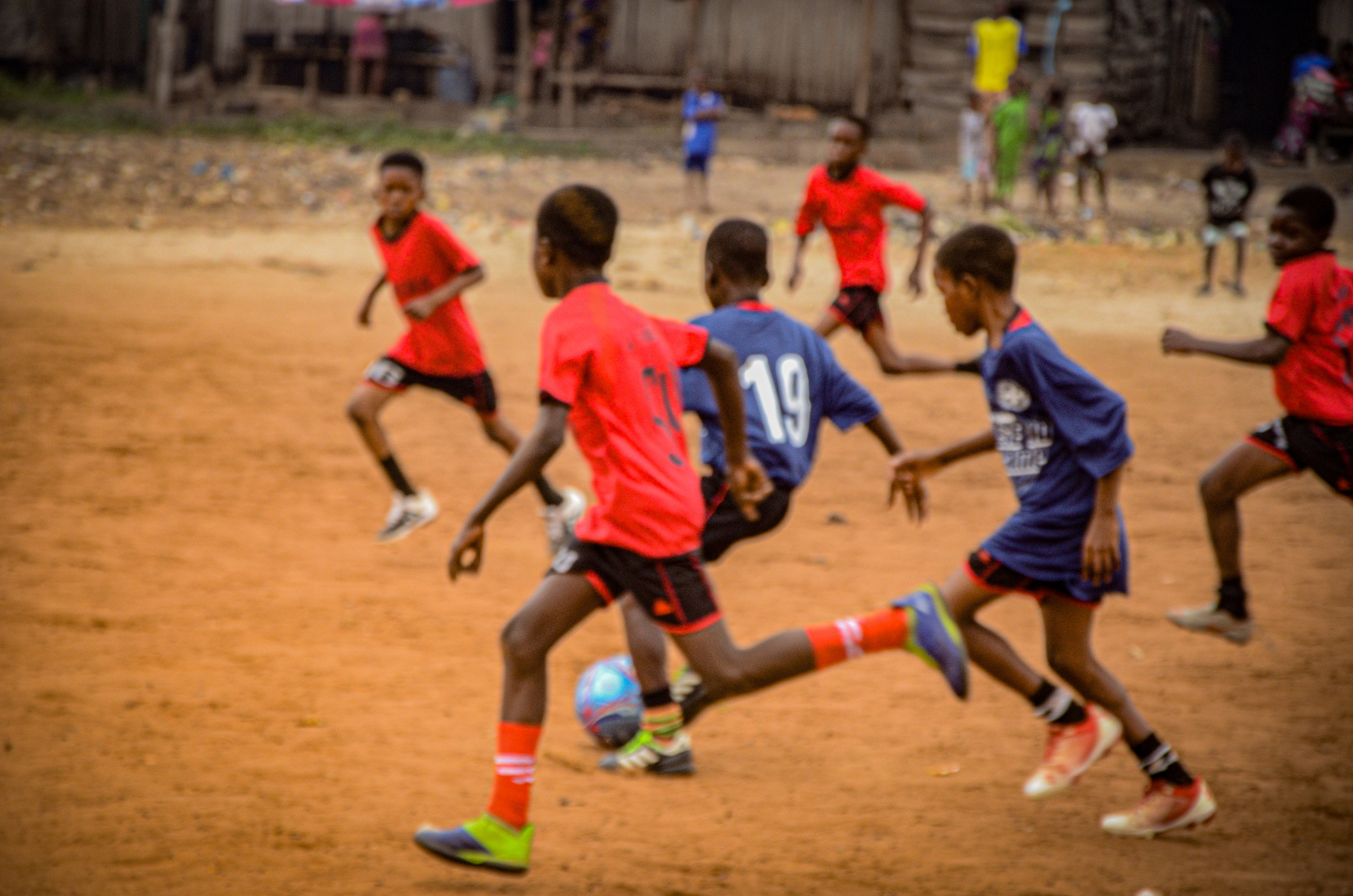 Children in coloured bibs playing football on a sandy pitch.