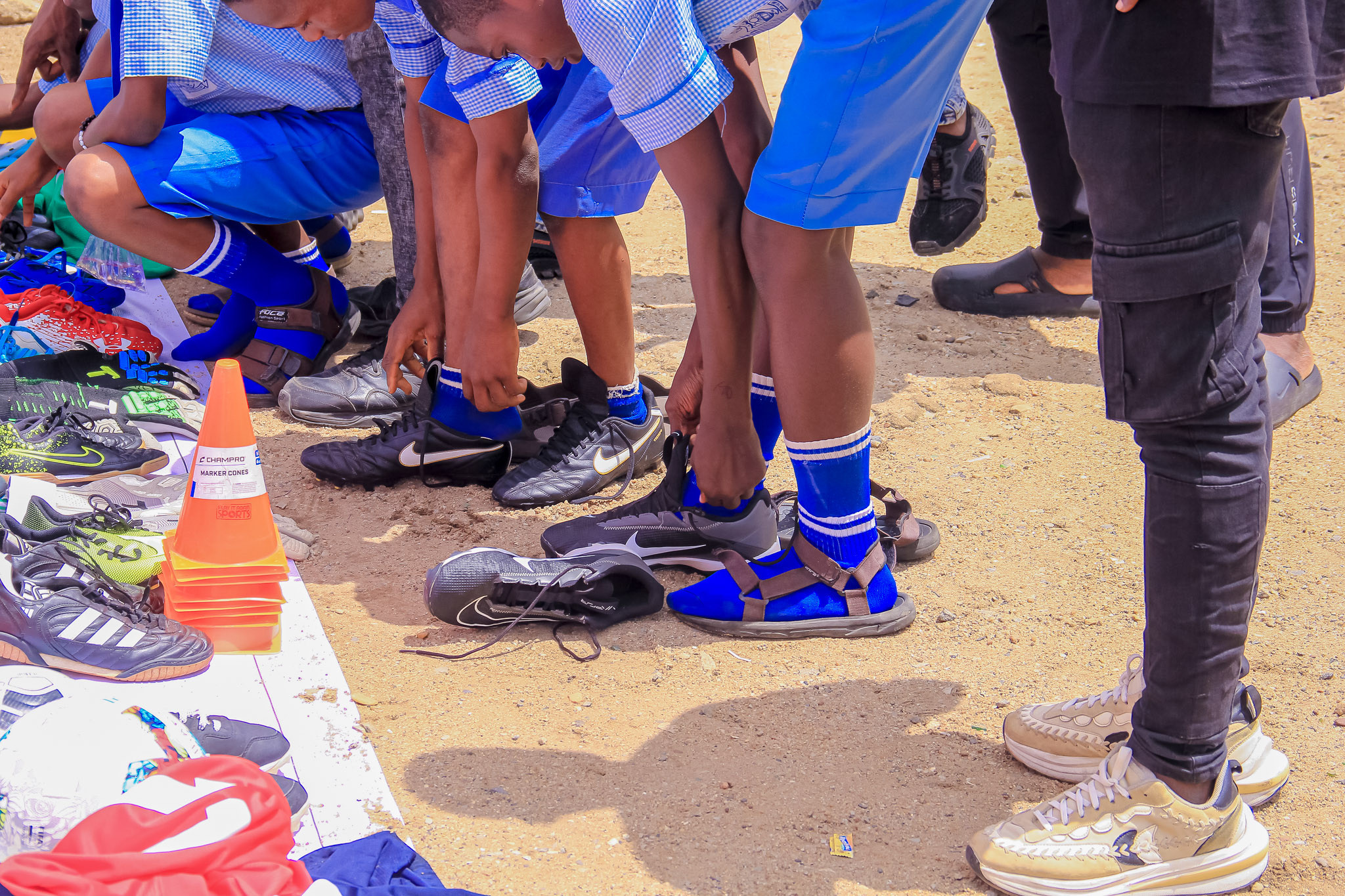 Young people crouching on a sandy pitch trying on football boots, with more boots laid out in front of them.