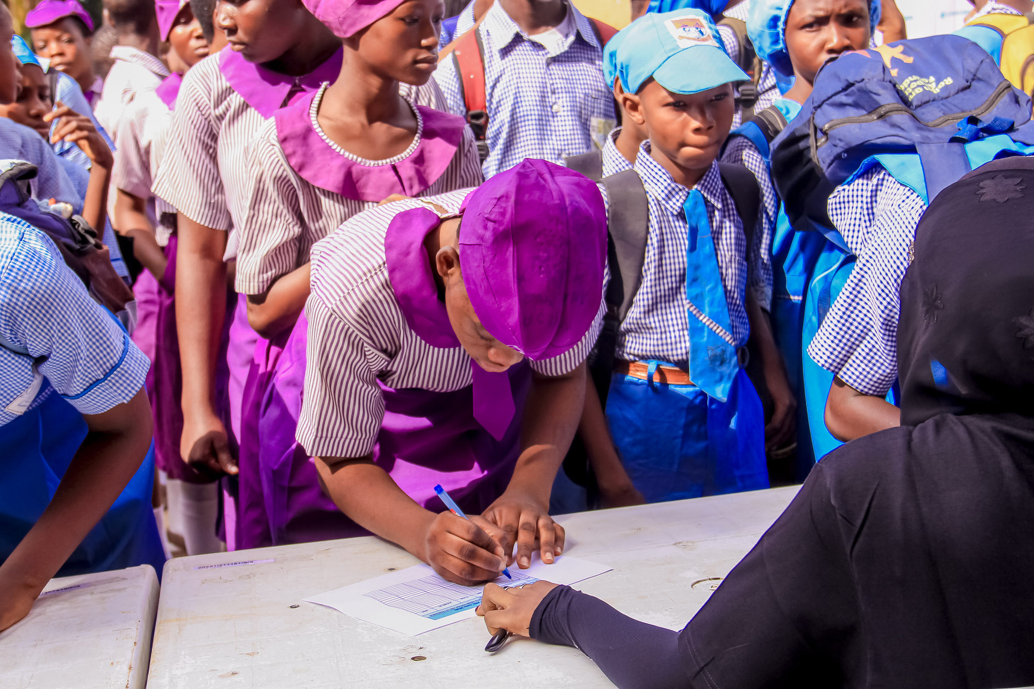 Students in school uniform crowded around a registration table while a volunteer writes on a form.