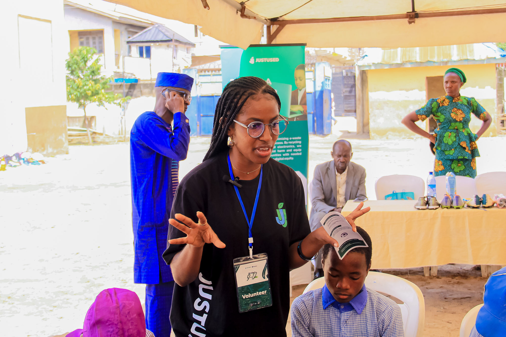 A volunteer in a JustUsedTech shirt speaking with a seated young person at an outdoor event.