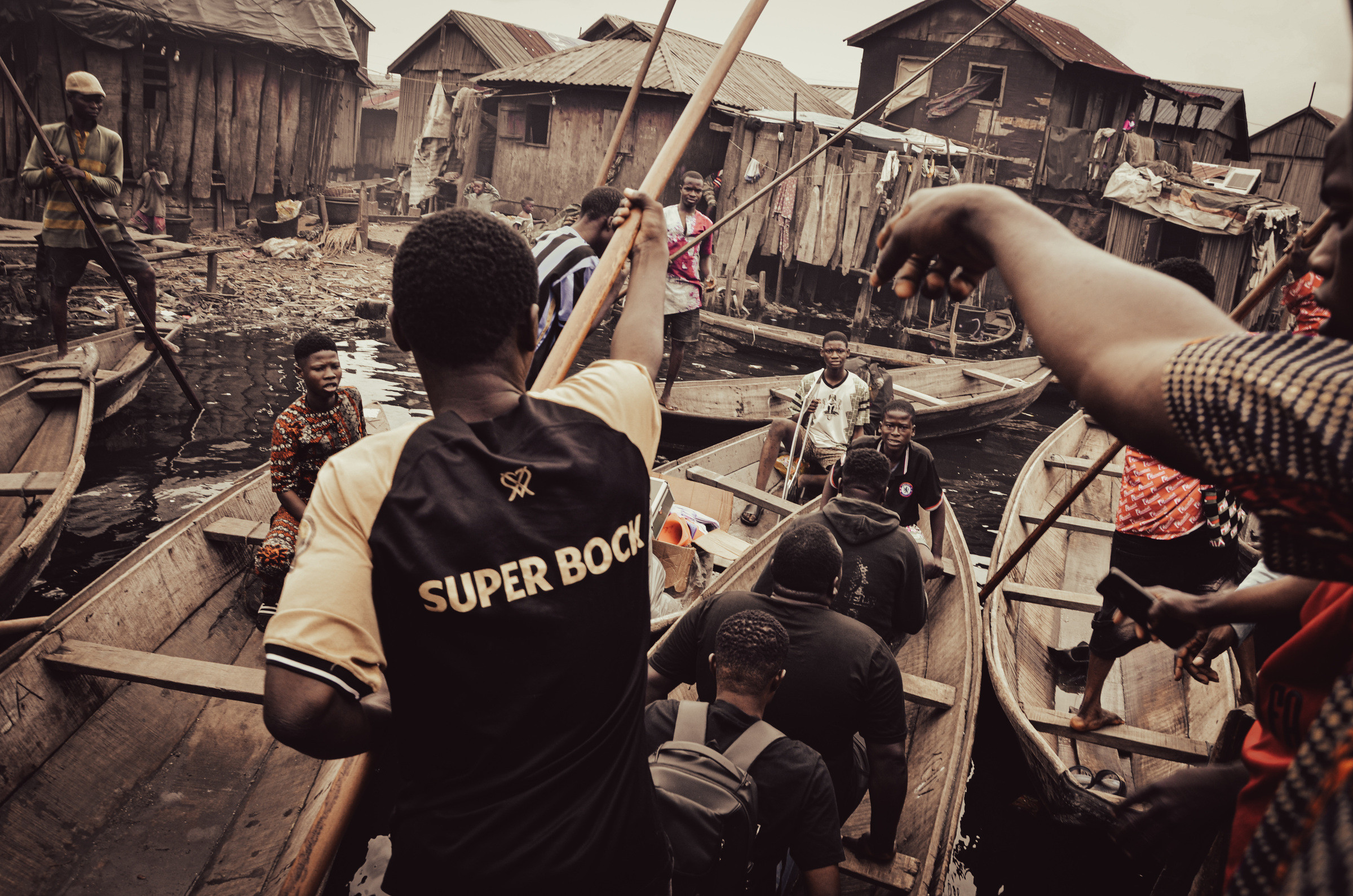 People moving between wooden canoes at a waterfront community, with stilt houses behind them.