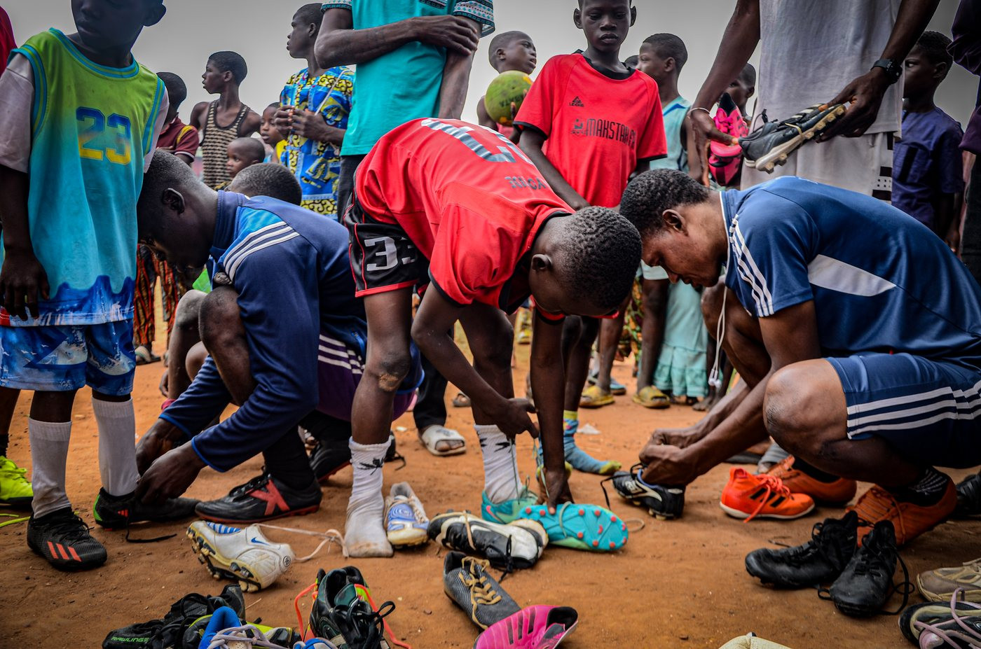 Young people crouching on a sandy pitch lacing football boots, with more boots laid out in front of them.