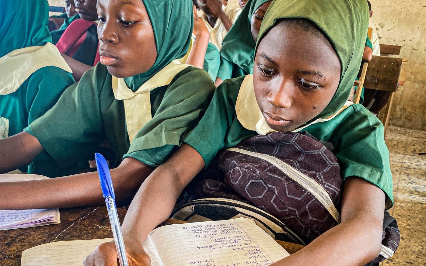 Two students in school uniform writing in an exercise book at a classroom desk.