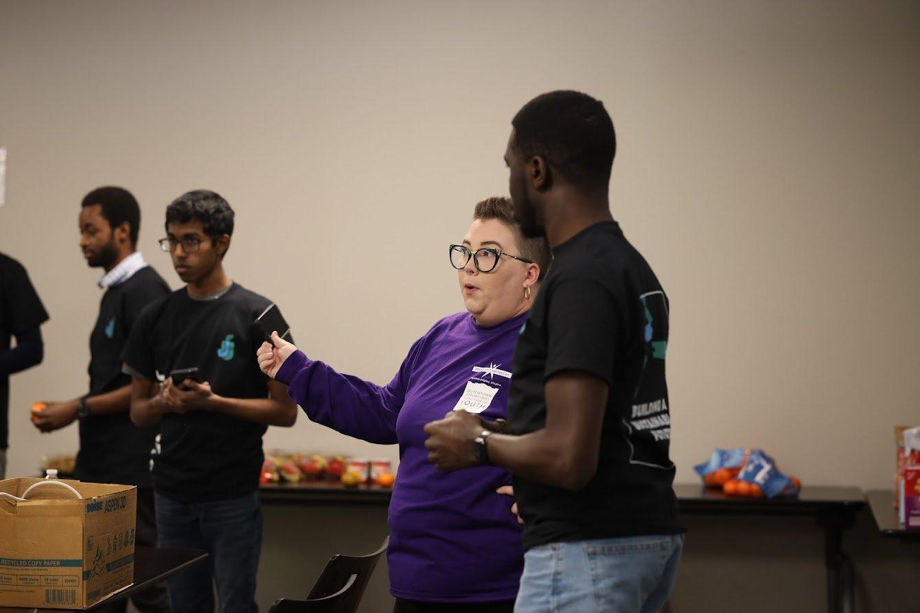 Two people in conversation at an indoor community event, with team members in JustUsedTech shirts and supplies laid out on tables behind them.