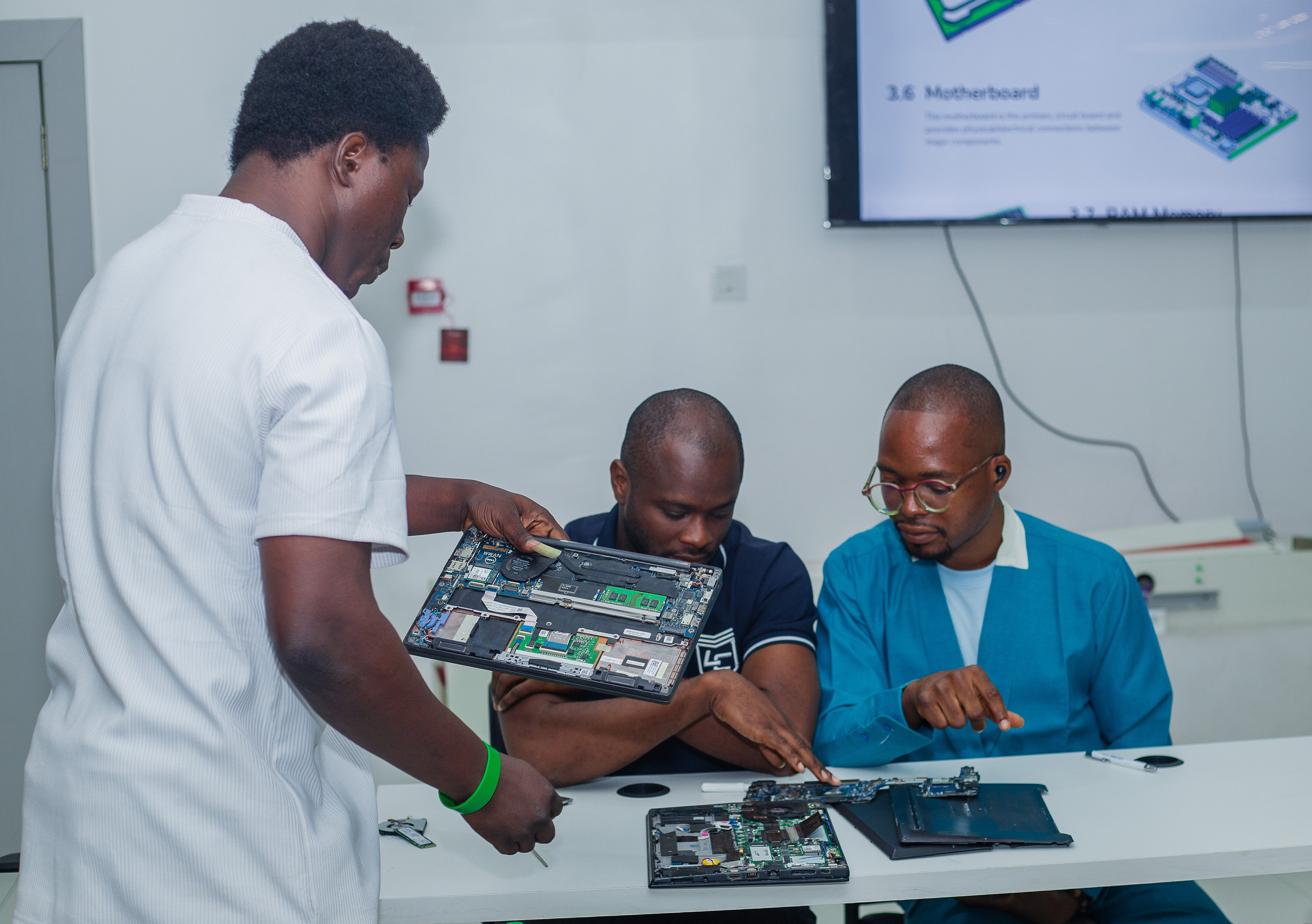 Three people examining an opened laptop, one holding the chassis up by its exposed board while the others study it, with a slide about motherboards on the screen behind them.