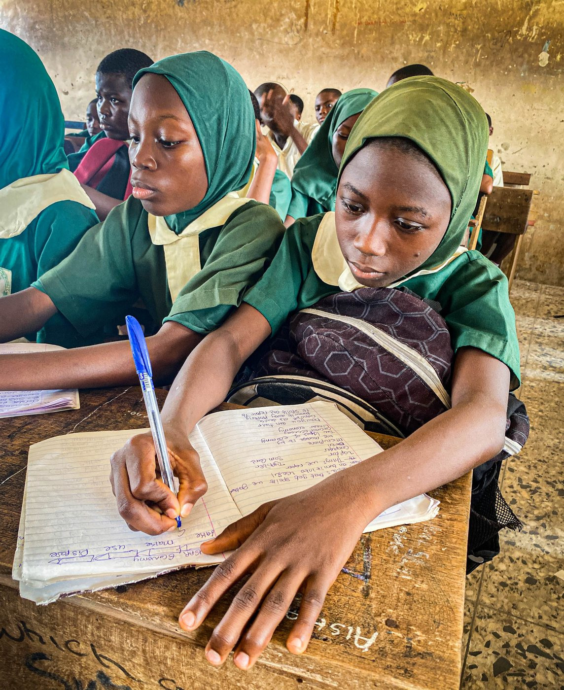Two students in school uniform writing in an exercise book at a classroom desk.